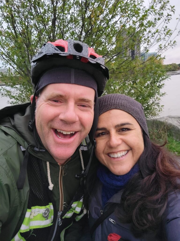John Richmond, wearing a bicycle helmet, poses beside his partner Paula Larrondo while they both smile